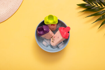 Homemade ice cream from molds on plate with ice on colored background and palm leaves, summer hat. Top view. Summer food background