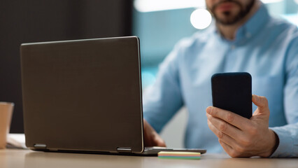 Unrecognizable mature businessman typing on smartphone, using laptop at office, selective focus. Banner with free space