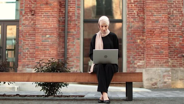 Cheerful young islamic businesswoman working on laptop while sitting near her office. Beautiful young woman with hijab working using laptop during break.