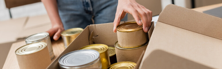 cropped view of african american volunteer packing canned food in box in charity storehouse, banner.