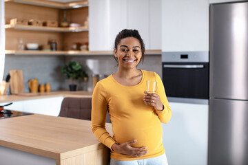 Happy young pregnant african american woman touching big belly and holding glass of water