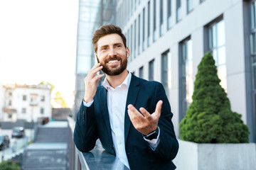 Happy confident european millennial bearded businessman in suit speaks by phone and gesticulates
