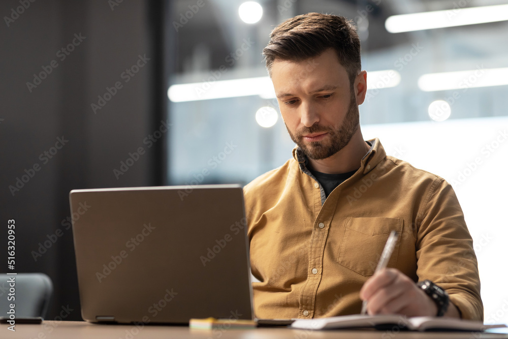 Poster serious businessman using laptop computer taking notes in modern office