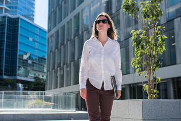 Young woman with laptop walking outdoors in business district. Woman commuting in office casual style. Portrait of a Businesswoman standing in a city district. 