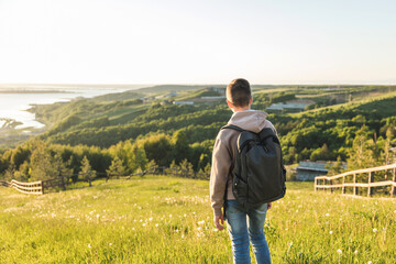 Tourist with backpack standing on top of hill in grass field and enjoying beautiful landscape view. Rear view of teenage boy hiker resting in nature. Active lifestyle. Concept of local travel © Lyubov
