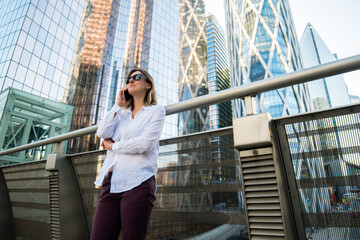 Young business woman in sunglasses  stands near office centre and talks on smart phone.  Business woman uses a mobile phone for work, calls. Business Woman With Phone Near Office. 