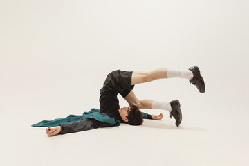 Portrait of stylish young man in black outfit and green coat posing, lying on floor isolated over grey studio background. Weirdness