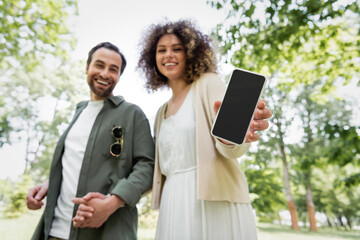 low angle view of happy curly woman holding smartphone with blank screen near smiling boyfriend in park.