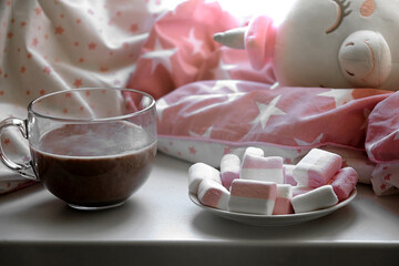 on the window on a child's pillow and under the blanket lies a pink unicorn children's toy next to a glass of cocoa and a plate with multi-colored marshmallows