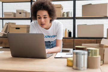 african american volunteer working on laptop near blurred tins in charity center.