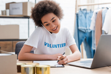 Fototapeta na wymiar smiling african american volunteer talking on smartphone and writing near laptop in charity center.