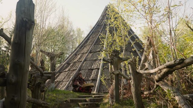 Musicain in Siberian shaman costume plays ritual drum sitting near ail building. Reconstruction of ancient Altai village on spring day