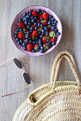 Bowl of strawberries and blueberries, sunglasses and wicker bag on the wooden table. Flat lay.
