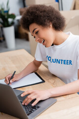 Fototapeta premium african american woman writing and talking on smartphone near laptop in volunteer center.