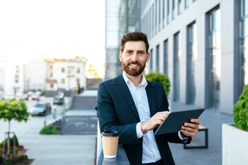 Fototapeta premium Smiling handsome confident european millennial bearded businessman in suit uses tablet to work