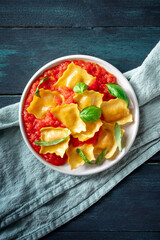 A plate of ravioli with tomato sauce and basil, top shot on a dark rustic wooden table, with a blue cloth