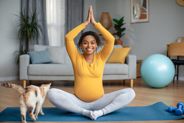 Cheerful young pregnant african american woman with big belly sits at lotus position, practices yoga
