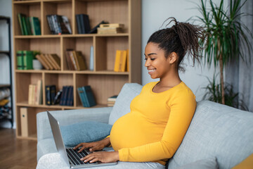 Cheerful millennial pregnant african american lady with big belly typing on laptop in living room...