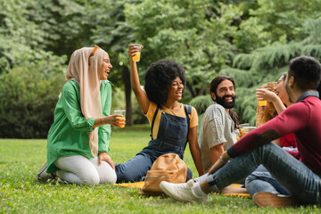 Naklejka premium group of multiracial friends picnicking in a park, african american woman raise glass with drink happy