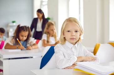 First grader elementary school. Portrait of cute little serious boy sitting at desk during lesson in school classroom. Handsome Caucasian blond kid boy sitting straight and looking at camera.