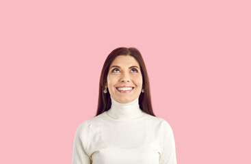 Happy young woman in white turtleneck standing isolated on pink background, looking up, smiling, dreaming of present, imagining something nice, thinking about holidays, choosing between two options