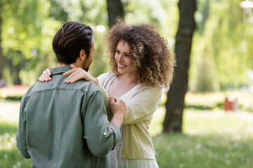 Fototapeta premium happy and curly woman in cardigan hugging boyfriend in green park.