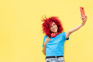 portrait of a smiling latin woman with red afro hair taking a selfie on a yellow background, wearing a blue t-shirt and red headphones around her neck.