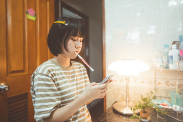 oung adult asian woman brushing teeth in font of bathroom mirror and typing on smartphone