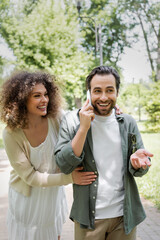 curly woman hugging cheerful boyfriend talking on smartphone in green park.