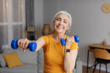 Positive senior woman doing dumbbell workout at home, working on arms strength and smiling at camera