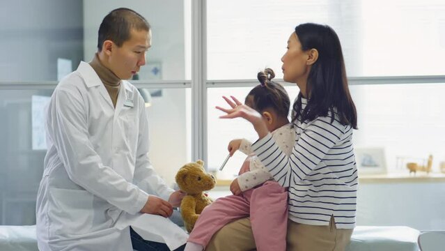 Asian mother sitting with toddler daughter on medial couch in clinic and having discussion with pediatrician
