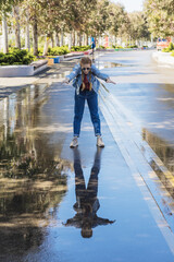 A woman in a denim outfit stands with her body tilted and her arms outstretched forward in a wet alley in a park. Walk after the rain. Mirror reflection in a puddle.