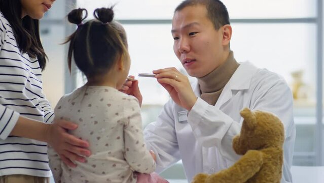 Asian pediatrician examining throat of toddler girl with flashlight while mother playing with her during medical checkup in clinic