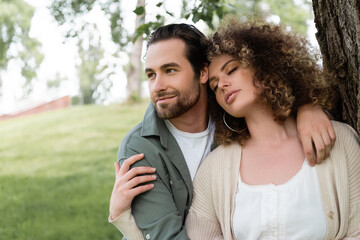happy man embracing curly and young woman with closed eyes near tree trunk.