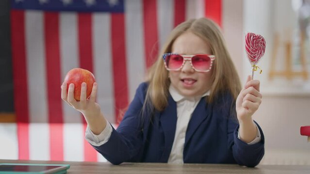 Healthful apple and unhealthy sweet lollipop in hands of blurred teenage schoolgirl choosing snack. Unsure teen girl sitting at desk in school classroom thinking with USA flag at background