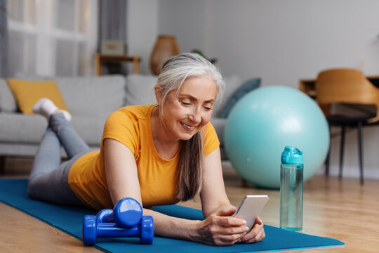 Senior woman resting on yoga mat, browsing internet on smartphone at home while taking break after home fitness - Powered by Adobe