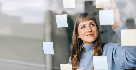 Young female entrepreneur sticking adhesive notes to a glass wall - obrazy, fototapety, plakaty