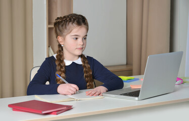 Cute girl in school uniform having video conference from home using laptop and writing in...