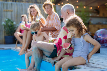 Multi generation family enjoy watermelon and drinks near backyard pool.