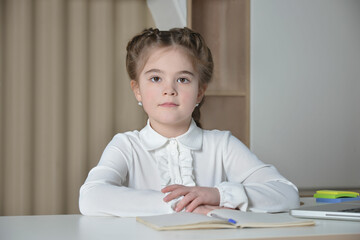 Naklejka premium A cute little girl from elementary school sitting at desk and looking at camera on a notebook, pen, multicolored paper clips stickers and curtains background.Back to school or education concept.