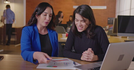 Senior business female executive looking at graphs and results with younger female staff at office...