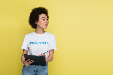 positive african american volunteer writing on clipboard and looking away isolated on yellow.