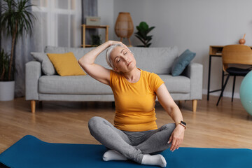 Healthy lifestyle concept. Senior woman stretching neck, sitting on yoga mat and warming up before...