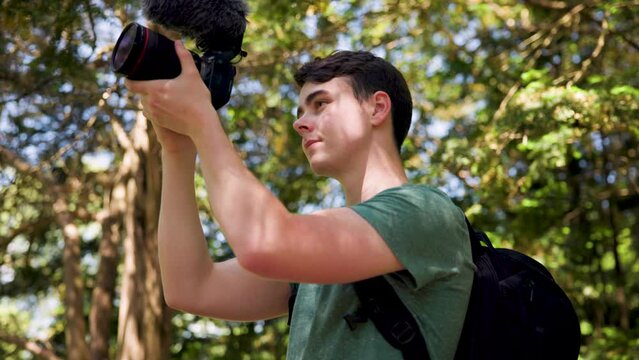 Close up of young male vlogger holding a professional camera shooting videos in a bright sunny forest