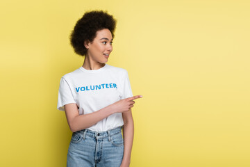 african american woman in t-shirt with volunteer lettering pointing with finger isolated on yellow.