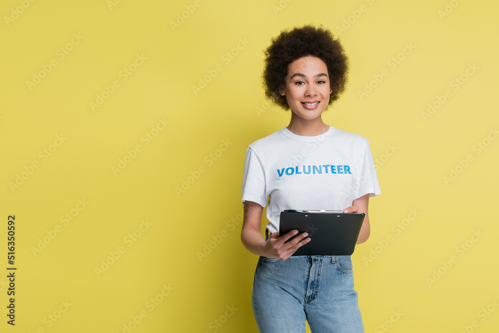 Wall mural african american volunteer with clipboard smiling at camera isolated on yellow.