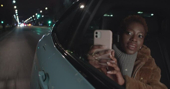 Positive smiling young black woman is taking photos with her cellphone through the car window in the night