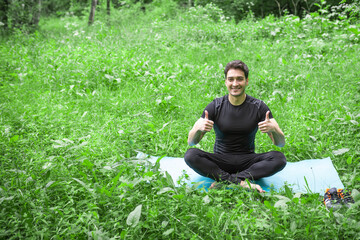 Naklejka na ściany i meble Cheerful sporty man in black sportswear sitting in lotus pose on fitness mat on the lawn and showing likes while doing exercise on nature close to forest
