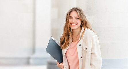 Young blonde student girl smiling against university. Cute girl student holds folders and notebooks...