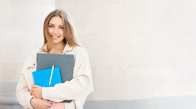 Young blonde student girl smiling against university. Cute girl student holds folders and notebooks in hands